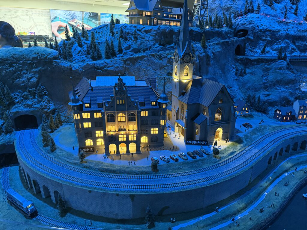 Night-lit miniature Swiss village with a town hall and church beside a curved railway, Nowon Train Village Swiss Hall