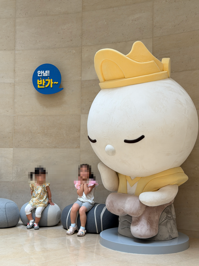 Two children sitting beside a giant soft sculpture of the Pensive Bodhisattva mascot in the museum lobby, Seoul