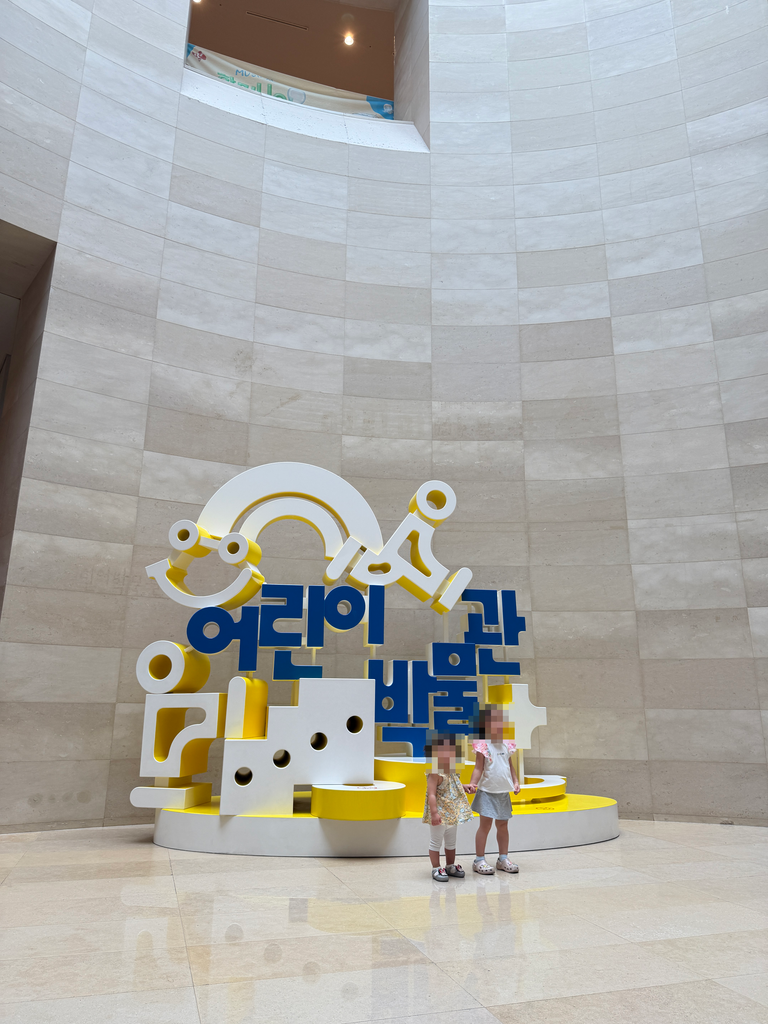 Children standing in front of the large white and yellow Children's Museum sign sculpture in the museum rotunda