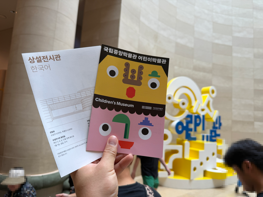 Hand holding a Children's Museum leaflet and a permanent exhibition guide in the museum lobby, Seoul