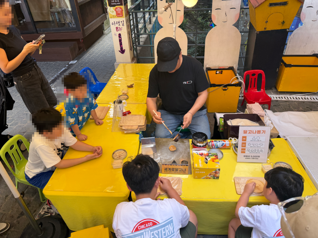 Children seated around a yellow table at a dalgona workshop in Insadong while the stallholder presses the melted sugar into a stamped shape