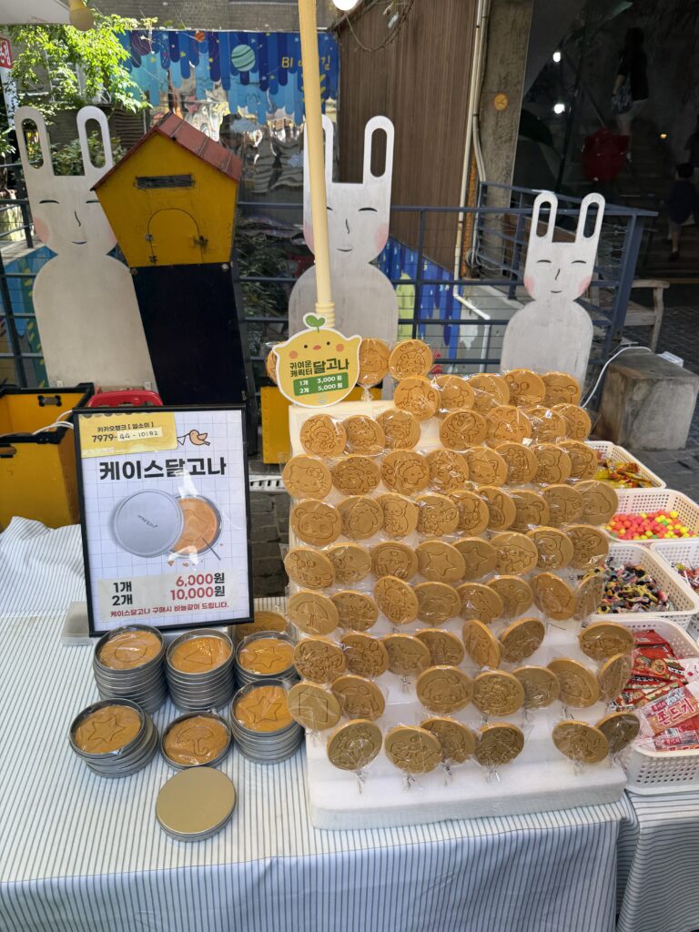 Display of dalgona honeycomb sugar candies in stamped shapes and tin cases at a stall in Insadong with prices shown