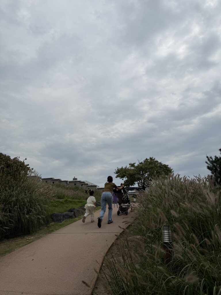 A woman pushing a buggy and a small child running along a paved path lined with pampas grass in Heunginjimun Park, Seoul