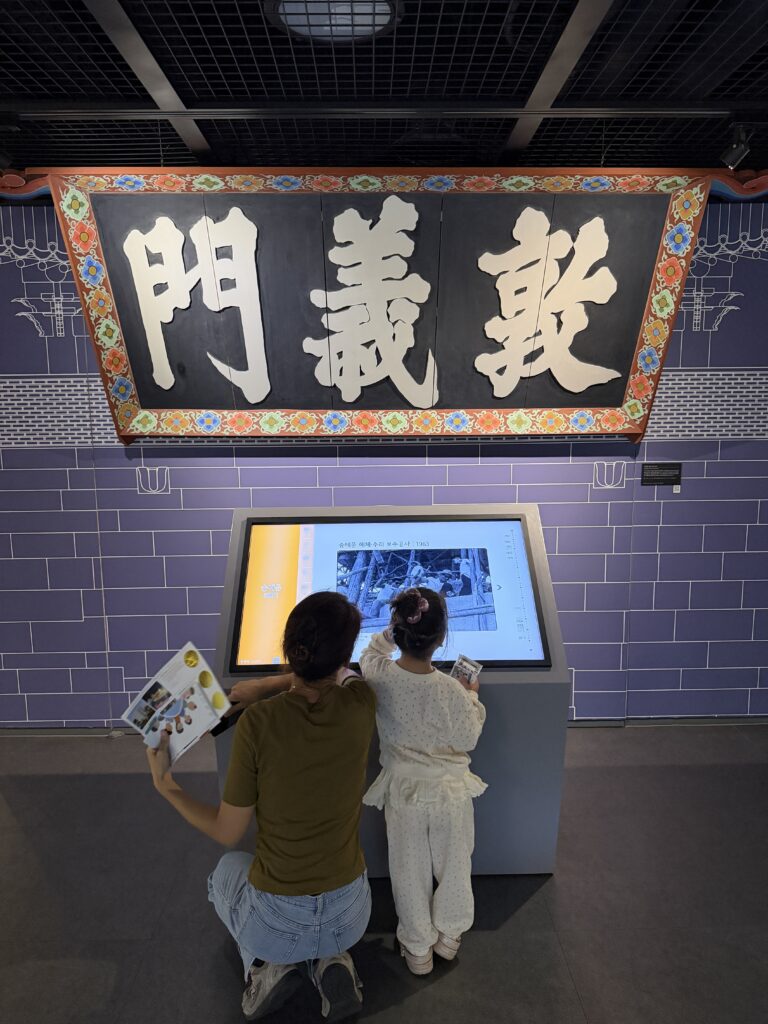 The wooden Donuimun gate plaque on display at the Seoul City Wall Museum with a visitor using the touchscreen below it