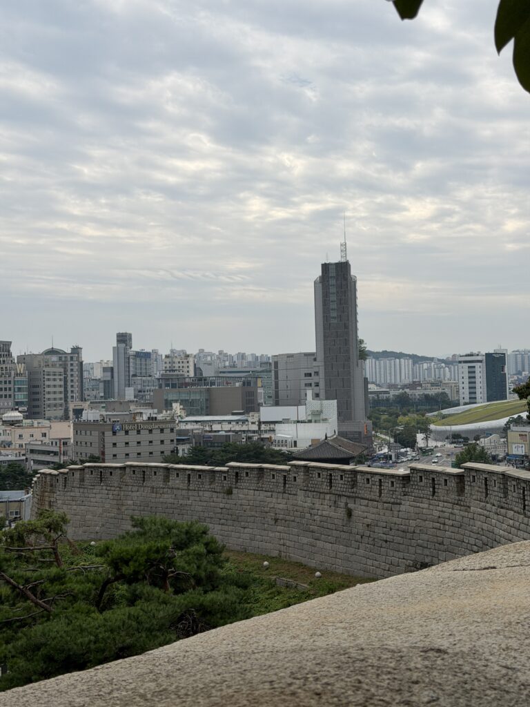 A dirt path running alongside a restored section of the Seoul City Wall under trees, with houses on the left