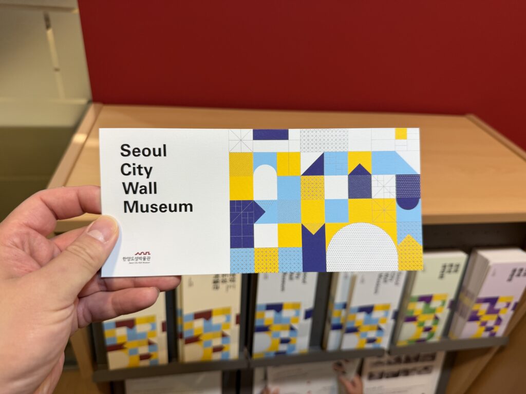 A visitor holding a Seoul City Wall Museum leaflet with a blue and yellow geometric design, in front of a rack of museum publications