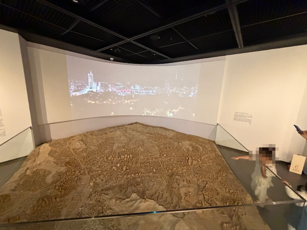 A large floor model of Seoul’s terrain at the Seoul City Wall Museum with a projected film of the modern skyline behind it and a small child pointing at the model