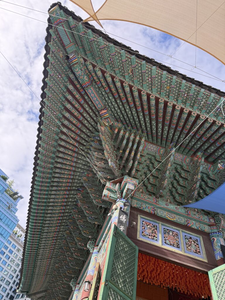 Upward view of the dancheong-painted eaves and rafters of the main hall at Jogyesa Temple, with a modern office building visible behind