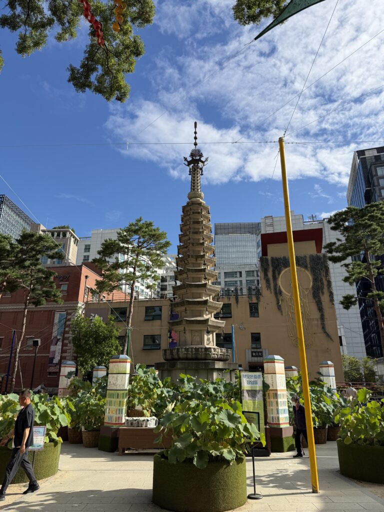  Octagonal ten-story stone pagoda at Jogyesa Temple surrounded by potted lotus plants, with modern office towers behind it