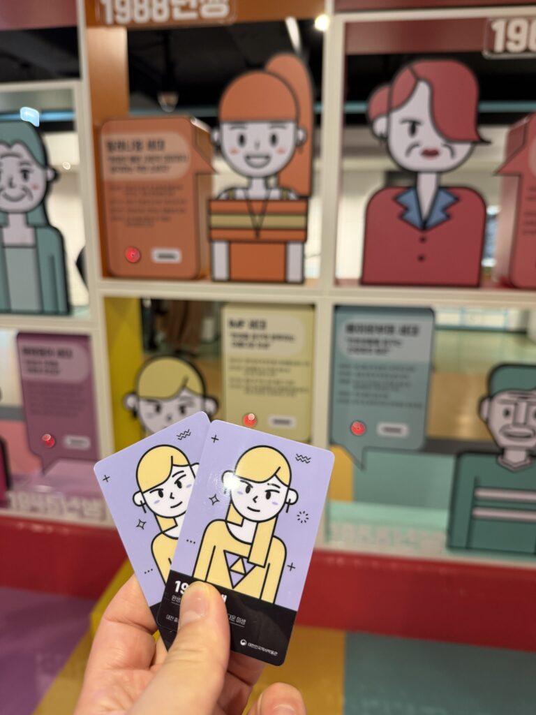 Two illustrated experience cards held up in front of a wall of cartoon characters from different decades of Korean history