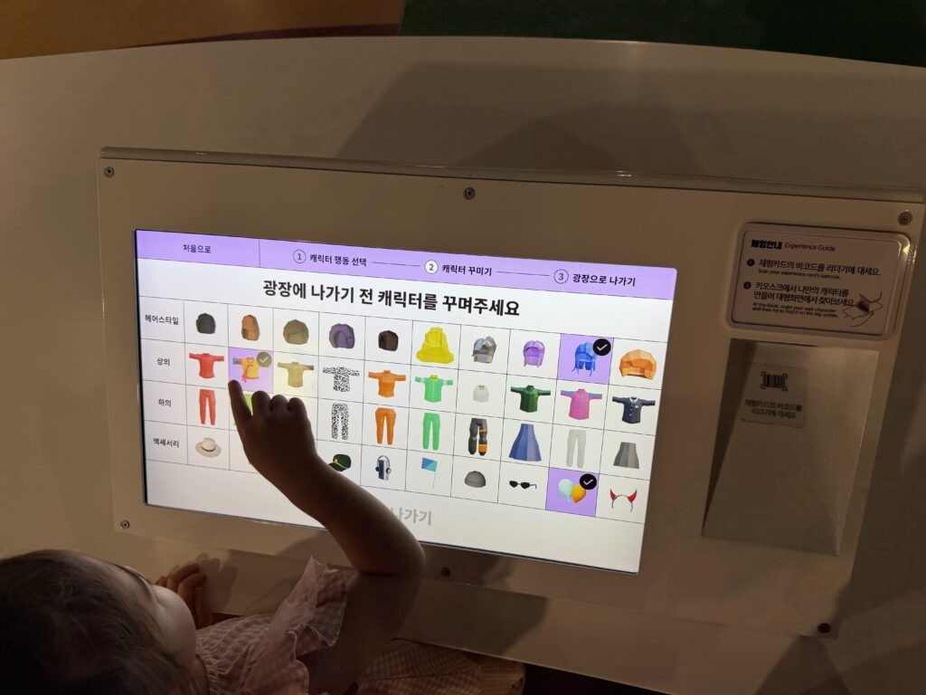 Child using a touchscreen kiosk to dress a character, with the barcode reader and bilingual guide panel to the right of the screen