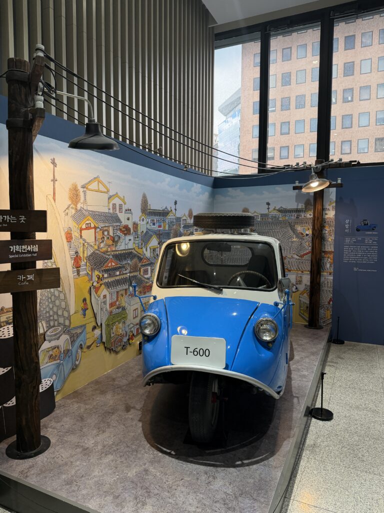 Blue T-600 three-wheeled truck from 1960s Korea displayed against a painted mural of a hillside neighbourhood