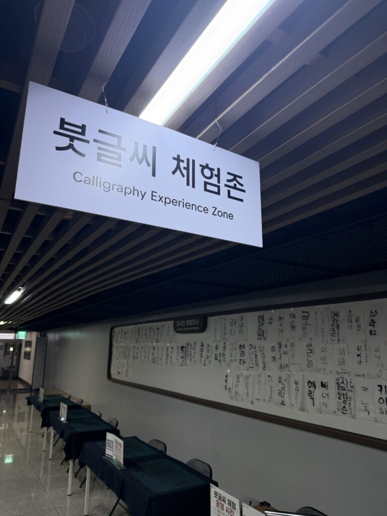 Calligraphy Experience Zone sign above desks with brushes and visitors' written sheets displayed on the wall behind_1