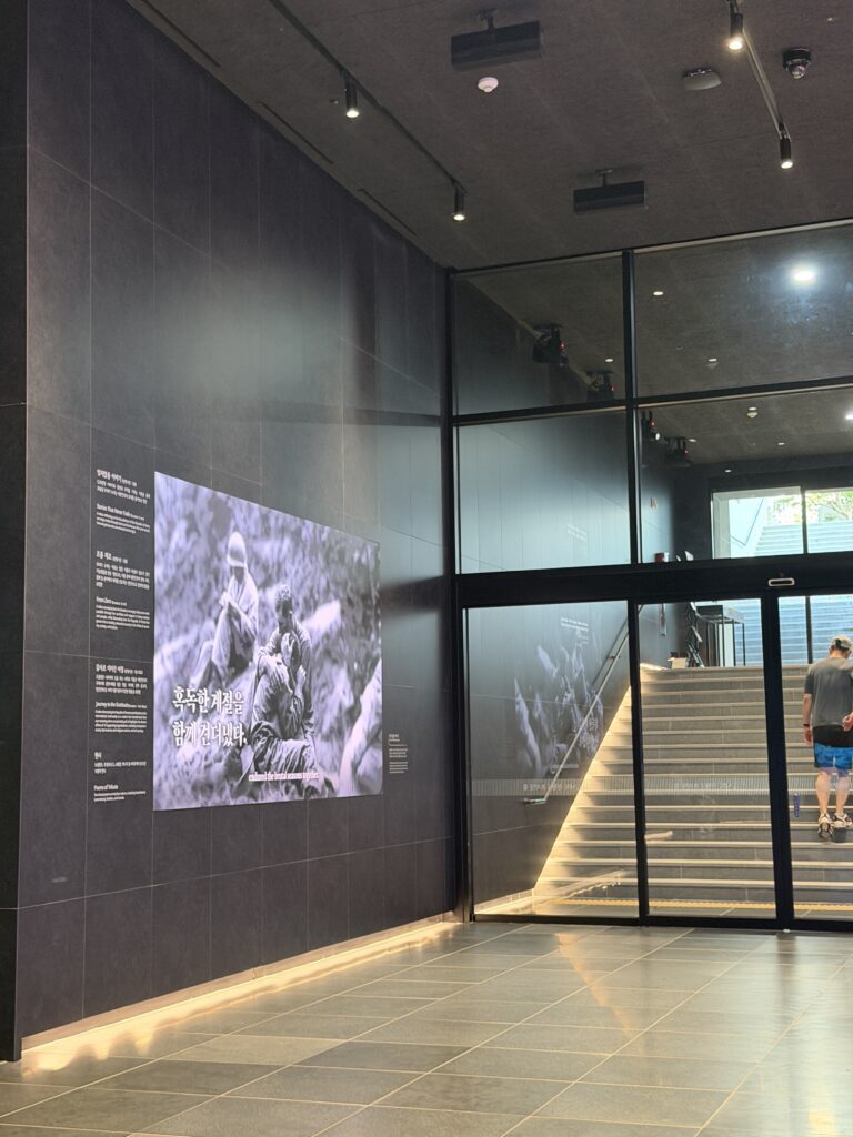 Dark stone lobby and staircase at the entrance to Freedom Hall beneath Gwanghwamun Square, with a Korean War photograph projected on the wall