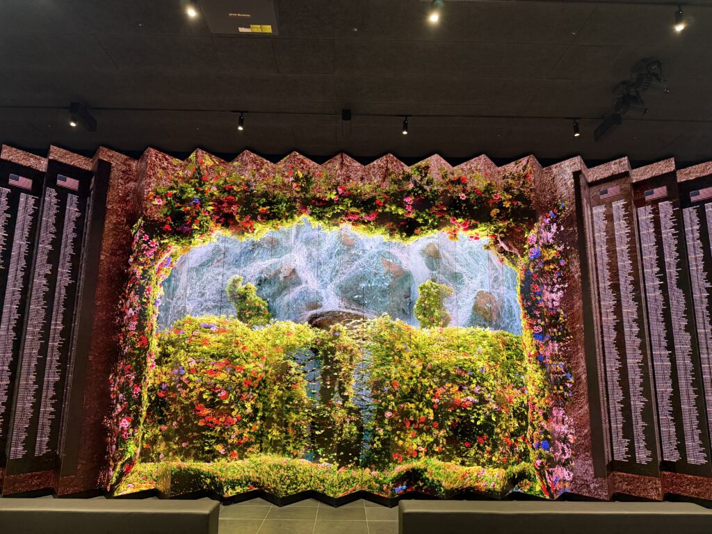 Memorial wall in Freedom Hall with columns of names on dark panels either side of a large media screen showing a flower garden